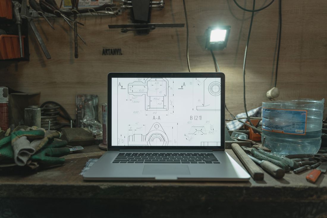 A laptop open on a cluttered workbench, plans on the screen.